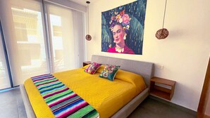 Une chambre à coucher avec un lit, des coussins colorés, une couverture jaune, un tableau accroché au mur et une petite table de chevet en bois.