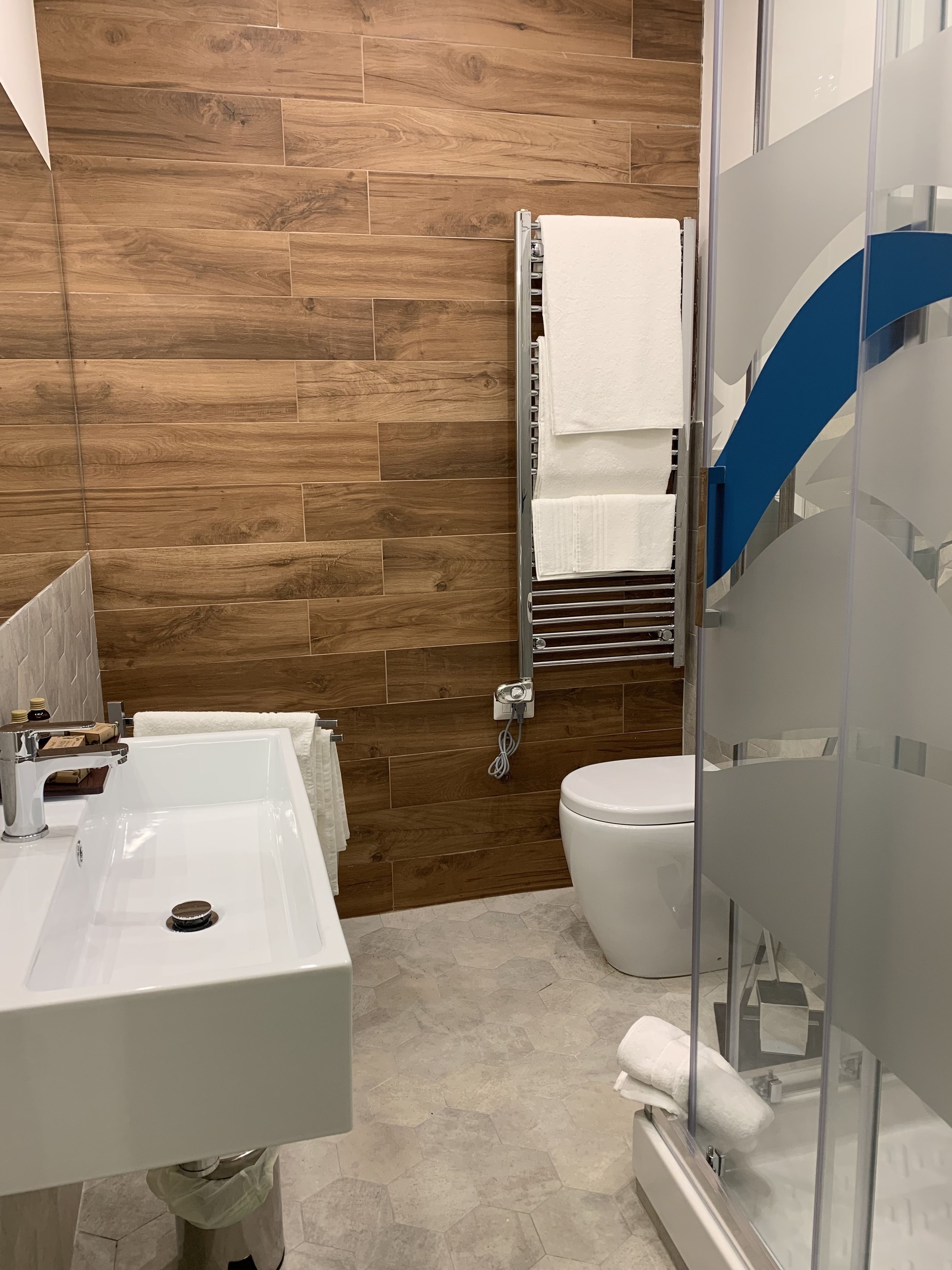 Un baño moderno con pared de madera, un lavamanos blanco, un inodoro y una ducha con mampara de vidrio.