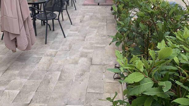 Un café al aire libre con mesas y sillas, suelo de piedra y vista al mar y un fuerte.