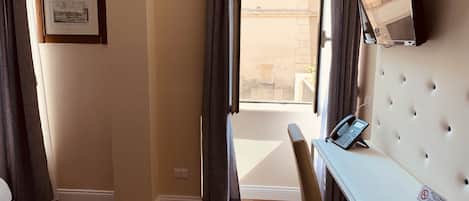 Habitación de hotel con piso de madera, cama blanca, escritorio con teléfono y ventana con cortinas.