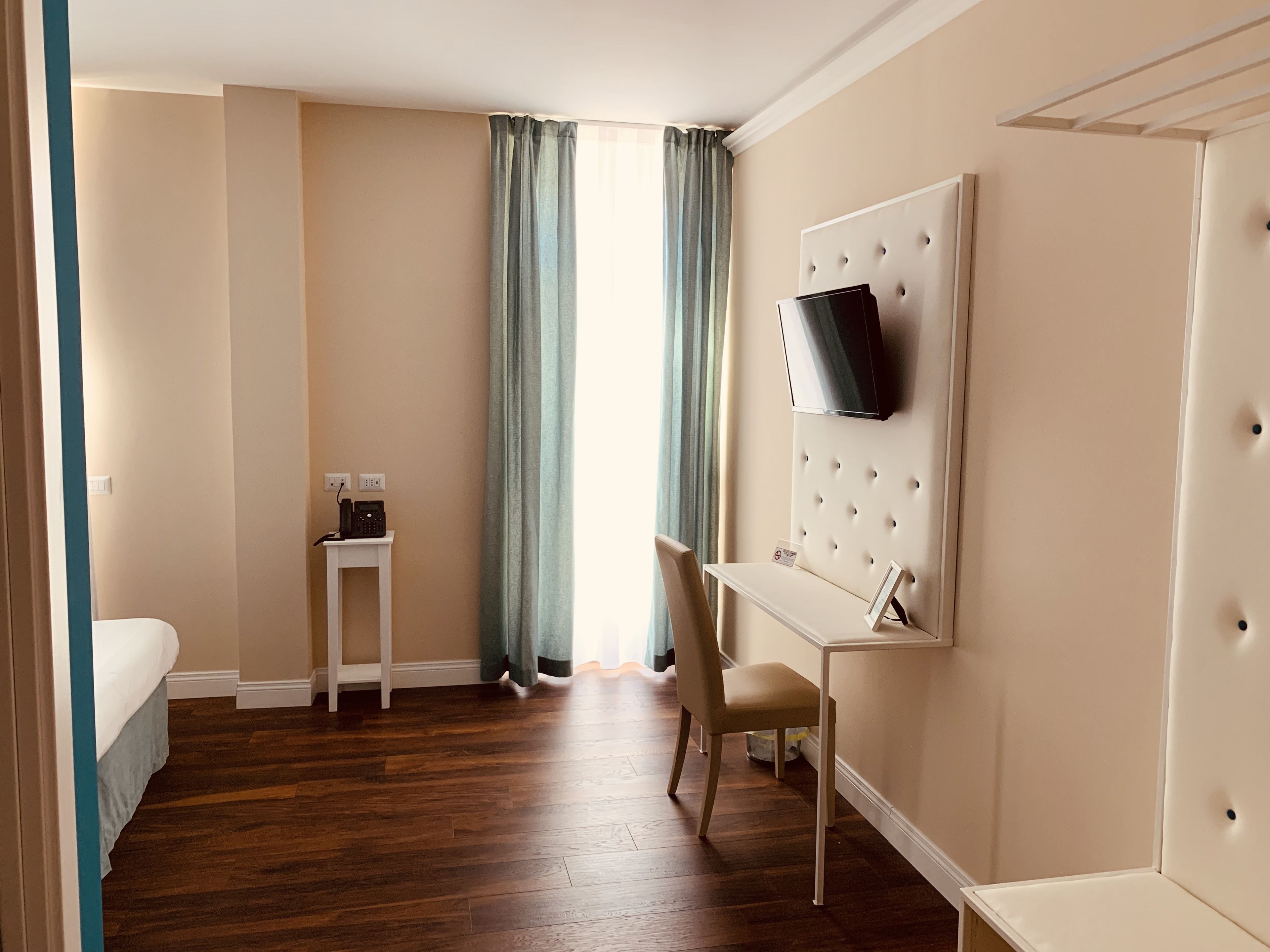 Habitación de hotel con cama, escritorio con silla, televisor montado en una pared acolchada y ventana con cortinas.