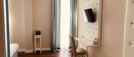 A hotel room with a bed, a desk with a chair, a television mounted on a padded wall, and a window with curtains.