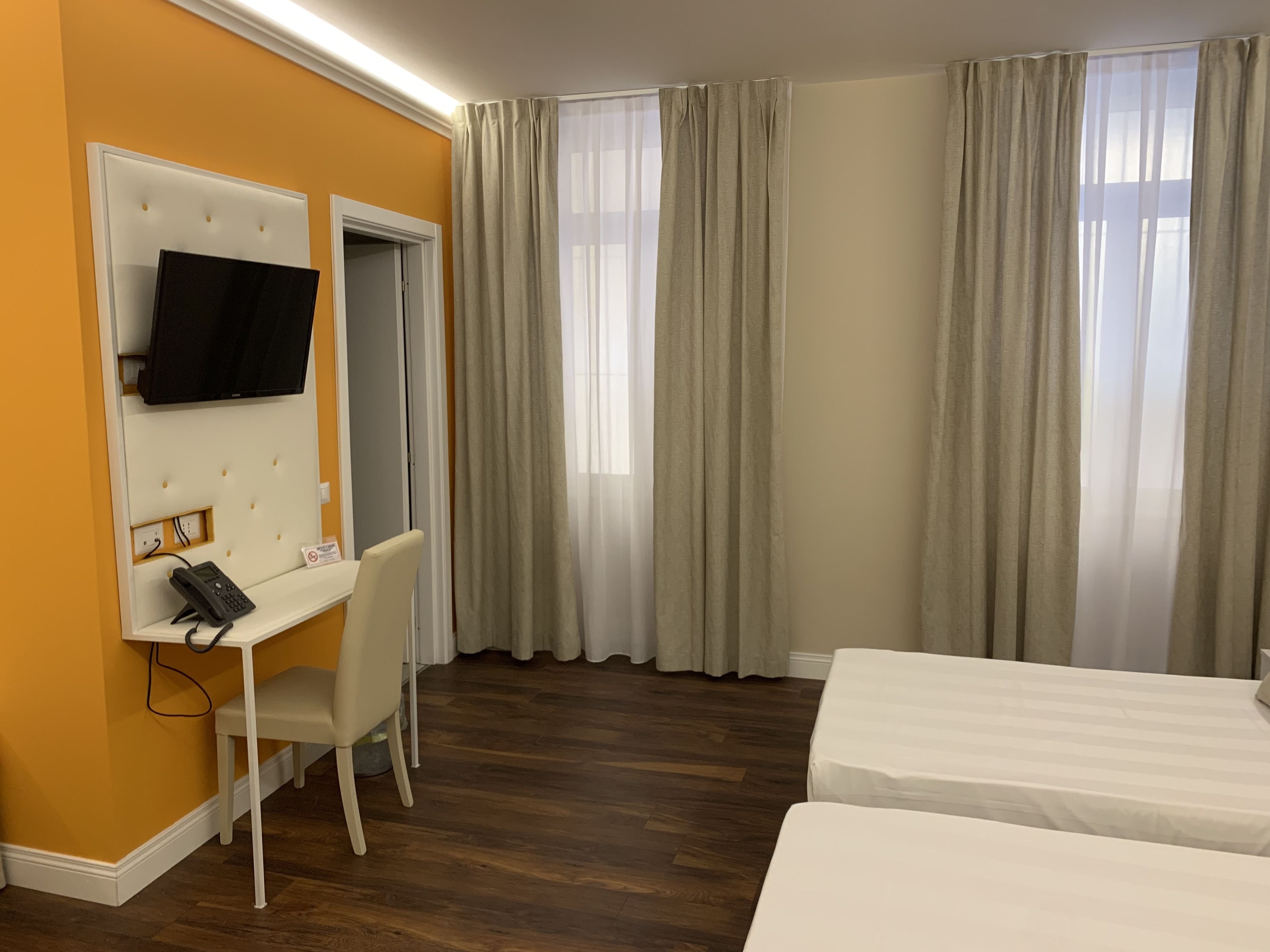Una habitación de hotel con cama, escritorio con silla, televisor montado en la pared y ventana con cortinas.