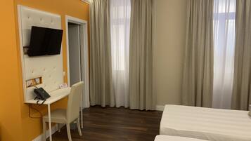 A hotel room with a bed, a desk with a chair, a TV mounted on the wall, and a window with curtains.