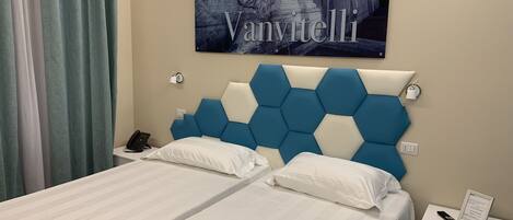 A hotel room with two beds, a blue and white headboard, a framed picture on the wall, a bedside table with a phone, and a nightstand with a lamp.