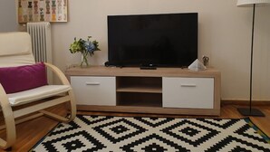 A living room with a TV on a wooden stand, a chair with a purple cushion, a floor lamp, and a patterned rug. - Mets Near From Panathenaic Kallimarmaro Stadium (Athens)