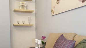 A cozy room with a beige sofa, a small black side table with two white wine glasses and a pink orchid, and a wall-mounted shelf with decorative items. - Mets Near From Panathenaic Kallimarmaro Stadium (Athens)