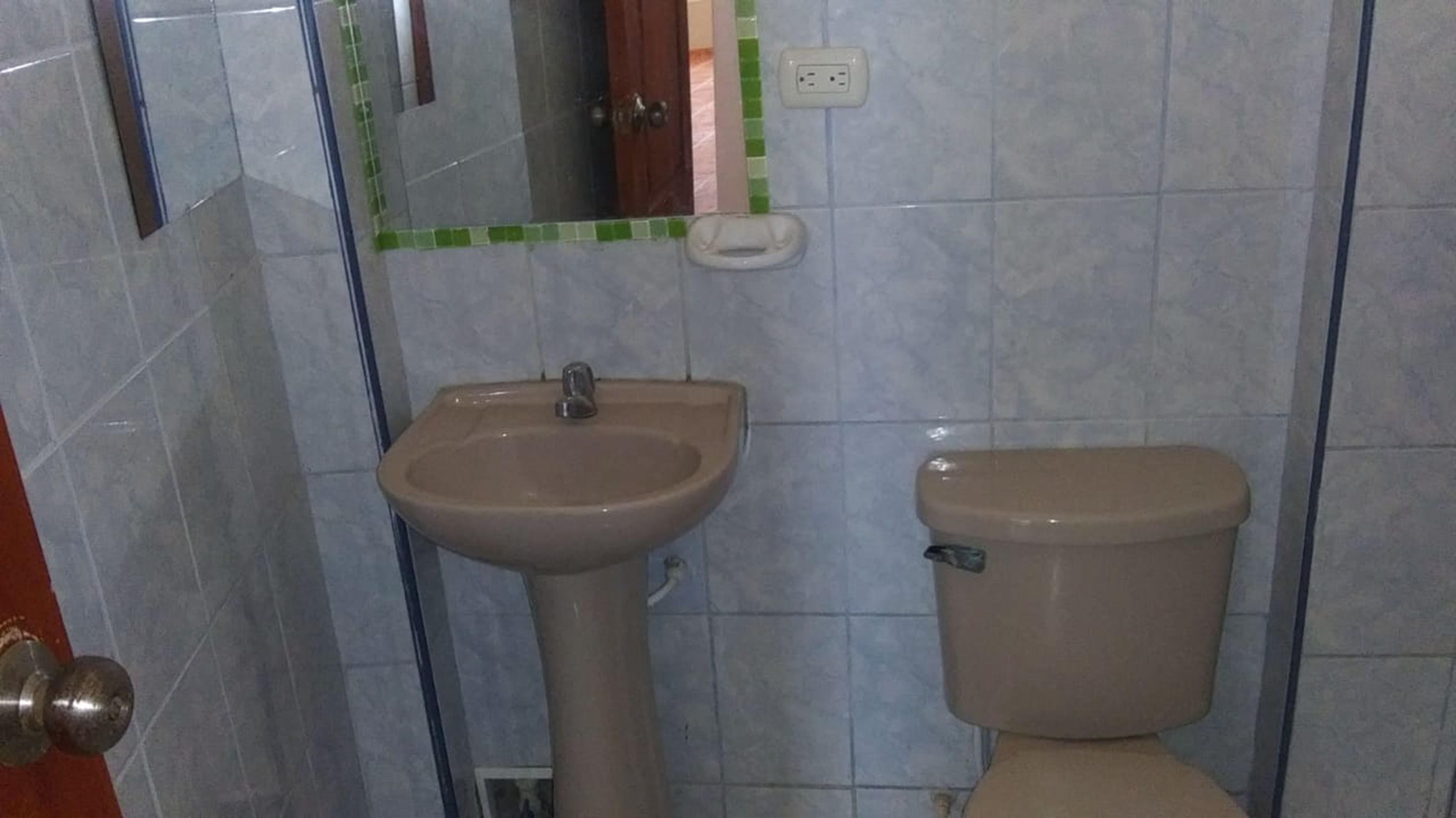 Un baño con un lavabo y un inodoro color beige, paredes con azulejos, un espejo con marco verde y un enchufe a la vista.