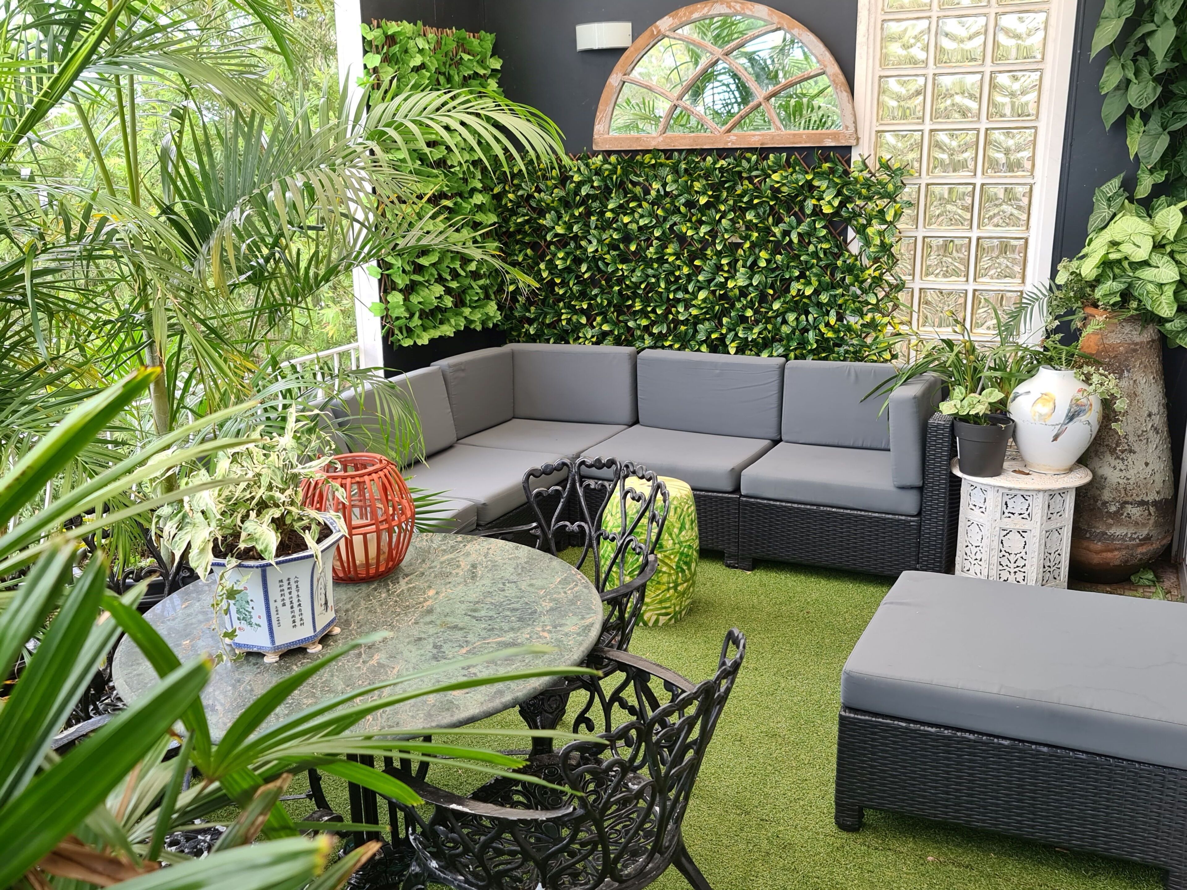 A cozy outdoor seating area with a curved gray sofa, a round table, and chairs, surrounded by lush greenery and a decorative wall.