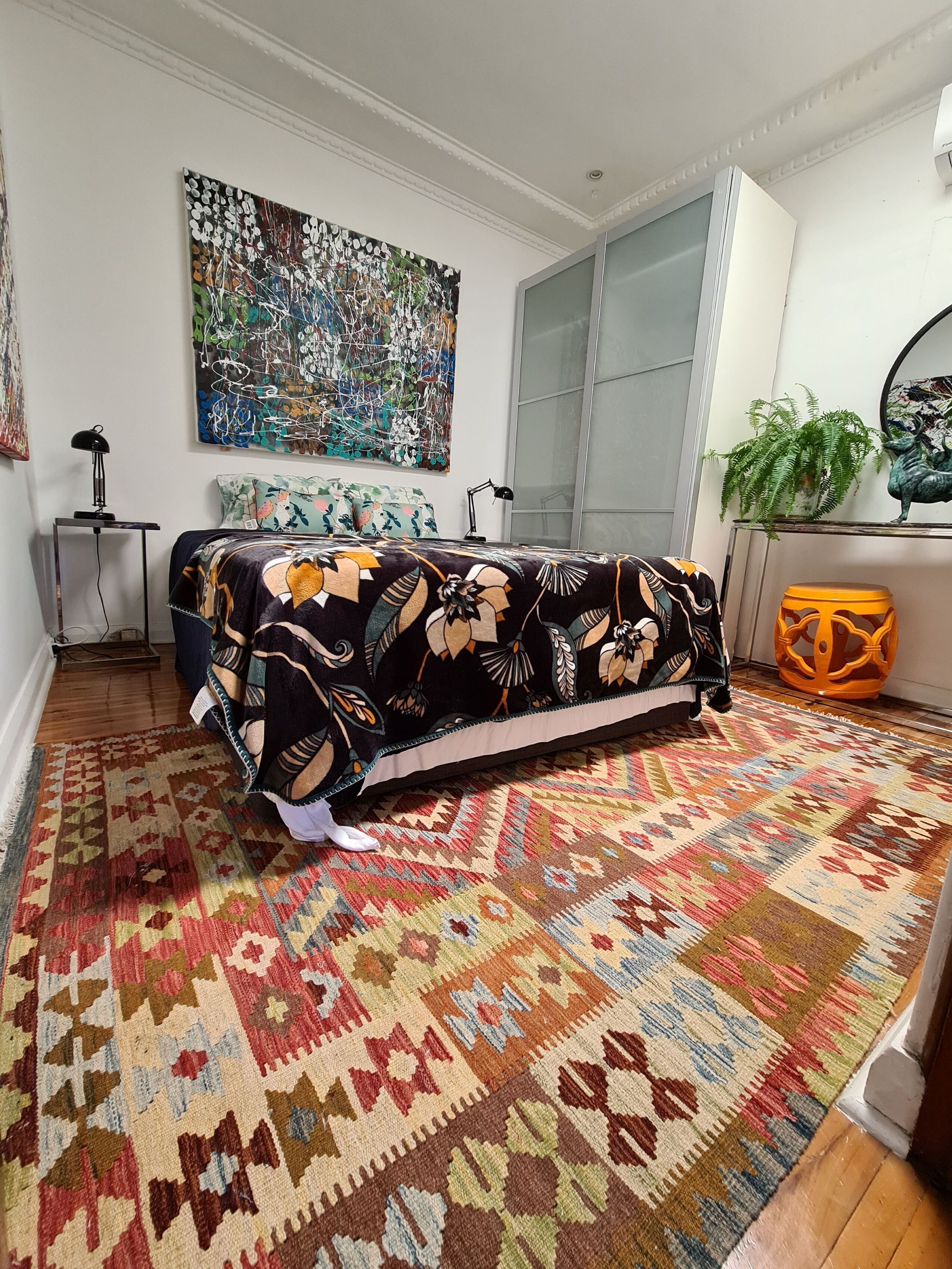 Une pièce avec un canapé recouvert d’une couverture à motifs, un grand tableau abstrait accroché au mur, une plante en pot et un tapis coloré.