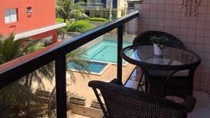 En balkong med flätade stolar och ett litet bord, med utsikt över en swimmingpool och de intilliggande byggnaderna.