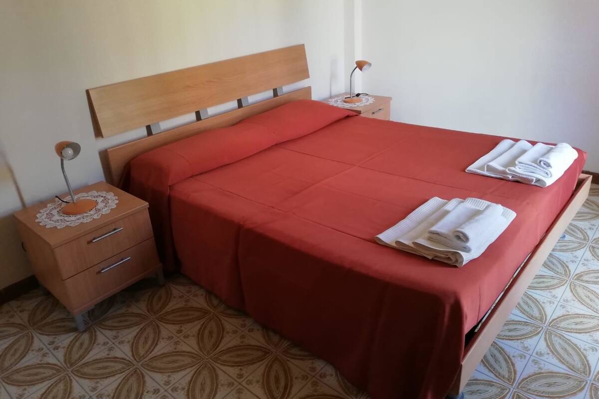 A neatly made bed with a red bedspread and two pillows, a wooden nightstand with a lamp, and a drawer.