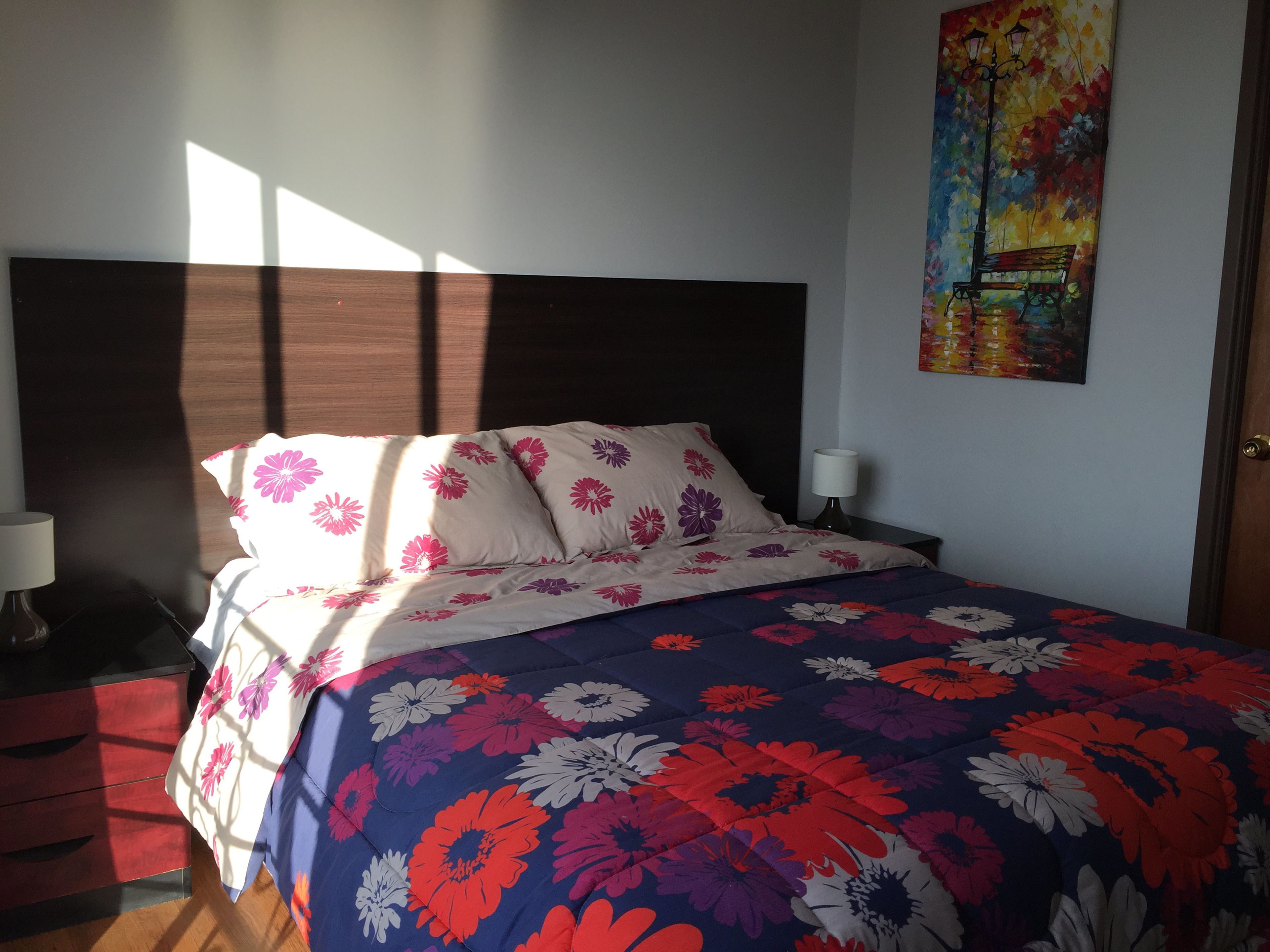 A bedroom with a bed, nightstands, a lamp, a painting, and a window with sunlight.