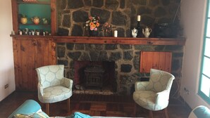 A cozy room with a stone fireplace, two patterned armchairs, a wooden bench, and a shelf with various items. - Yemanya Hostal (Concon)