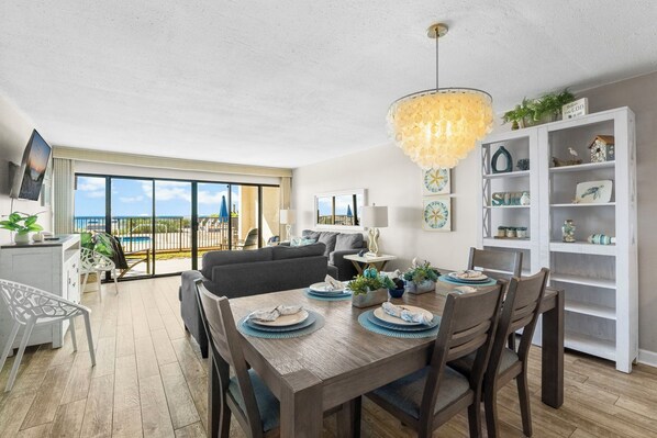 A spacious living room with a dining area, large windows with a view of the ocean, and a white shelving unit with decorative items.