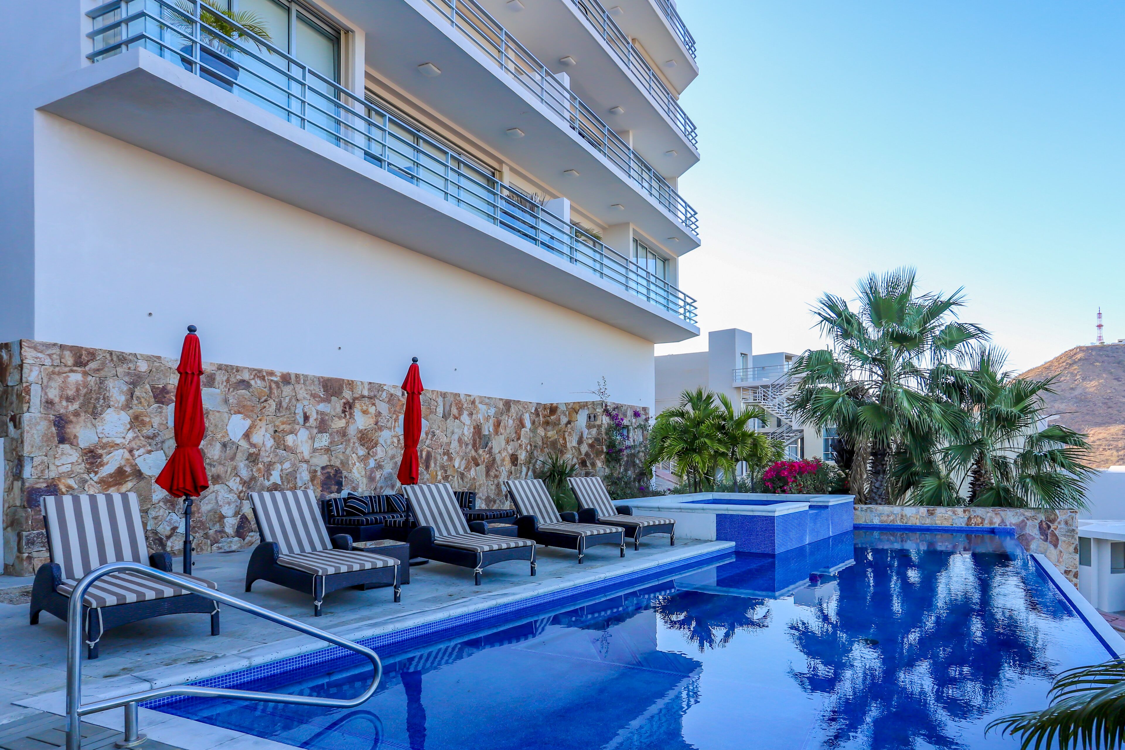 Zona de piscina con sillones de playa y sombrillas, un muro de piedra y vista a un edificio con balcones.
