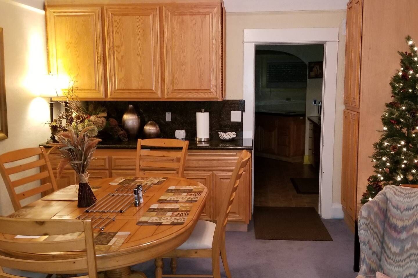 A dining area with a wooden table, chairs, and a vase of dried plants. There is a kitchen visible through an open doorway, and a Christmas tree with lights is in the corner.