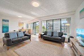 A modern living room with two black leather sofas, a large flat-screen TV, and a view of the city through large windows.