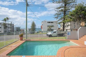 A swimming pool with a brick border, surrounded by a white fence, and a view of a residential area with buildings and a road.
