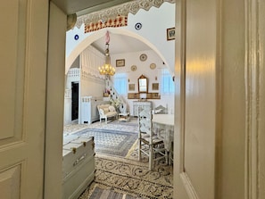 A room with a white arched doorway, a chandelier, a white sofa, a bed with a white headboard, and a patterned rug.