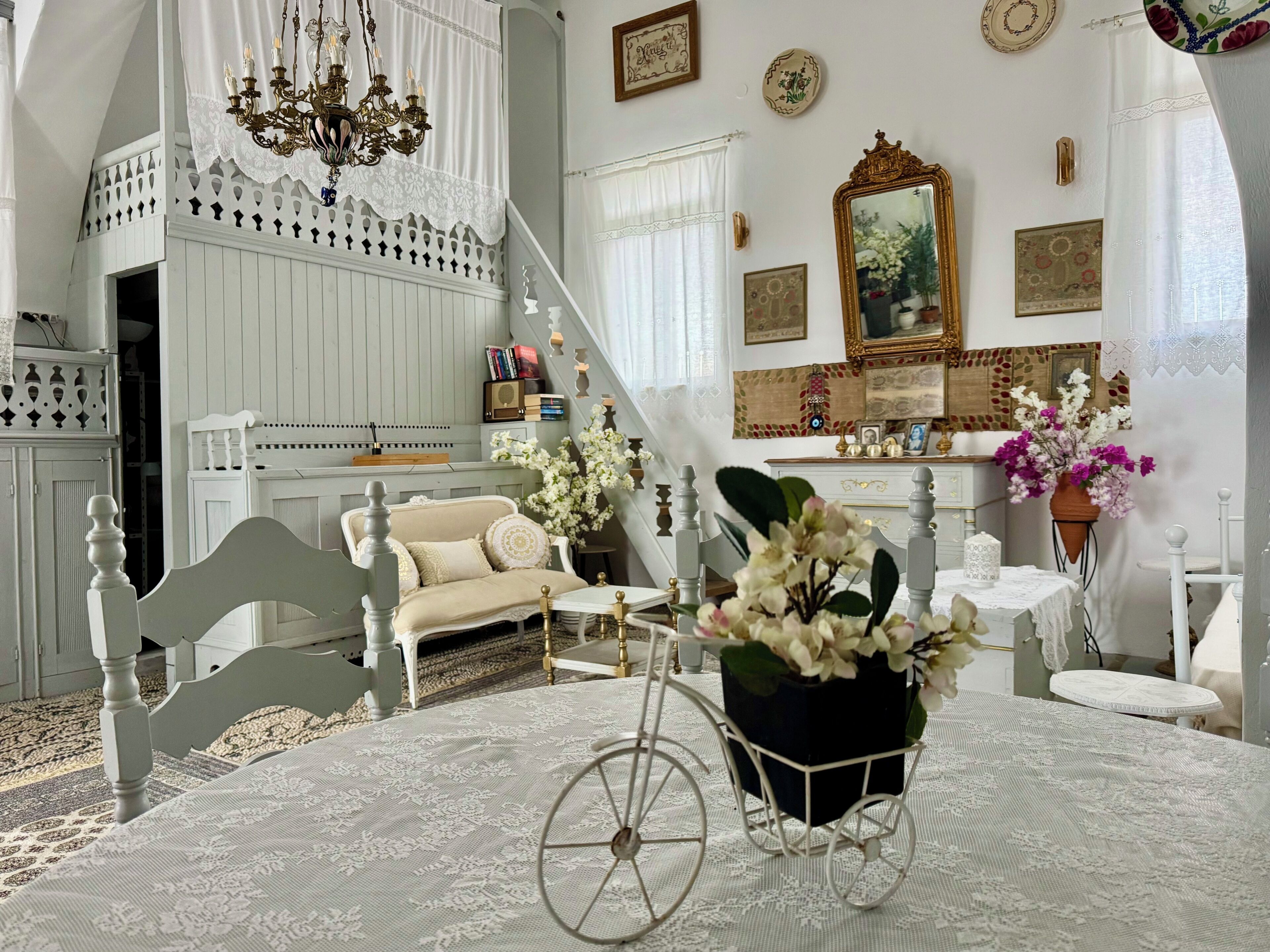 A room with a white sofa, a bicycle cart with flowers, a chandelier, and a staircase.