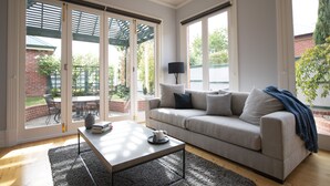 A living room with a sofa, coffee table, and a view of a brick building and greenery outside. - Charming Historic Home With Modern Interior - In The Heart Of Ballarat (Ballarat Central)