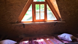 A cozy attic bedroom with a sloped ceiling, a wooden window with a view of greenery, and a colorful striped rug.