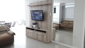Un salon moderne avec un téléviseur à écran plat fixé sur un meuble en bois, un canapé beige et une cloison en verre qui sépare l’espace de vie d’une chambre à coucher.