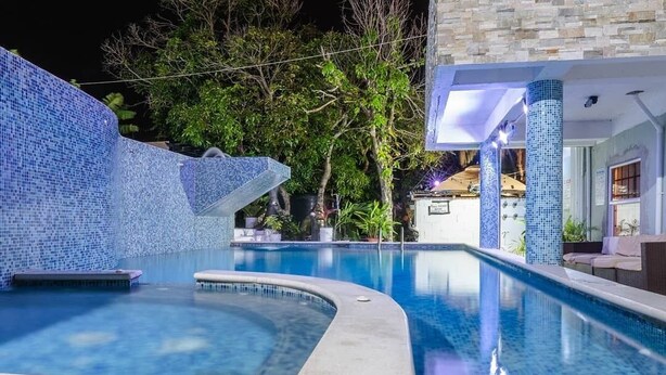 Una piscina con un muro de mosaico, rodeada de vegetación y un edificio con pilares azulejados.