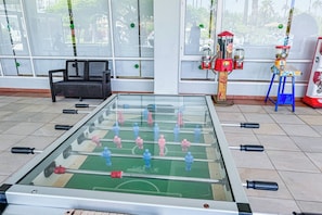 Een tafelvoetbaltafel in een kamer met grote ramen, een bank en een oude frisdrankdispenser.
