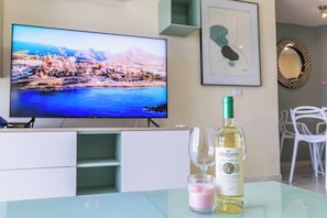 Een moderne woonkamer met een flatscreen televisie waarop een pittoresk stadsgezicht aan de kust te zien is, een fles wijn en twee glazen op een glazen tafel.