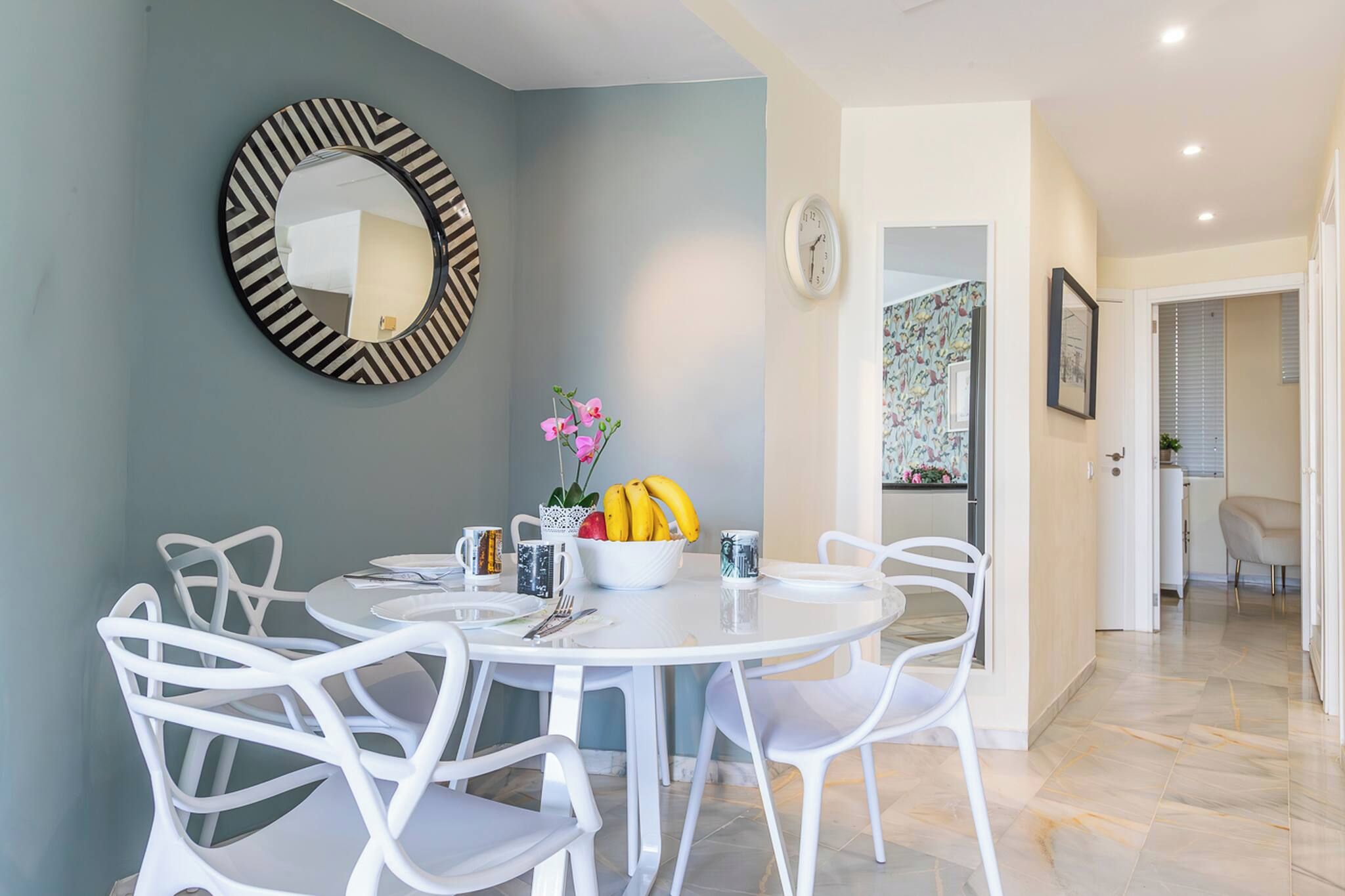A dining area with a round table, white chairs, a bowl of fruit, and a decorative mirror on the wall.