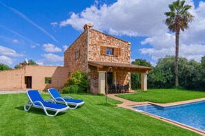 Una casa de piedra con piscina, dos sillones de descanso y una palmera.