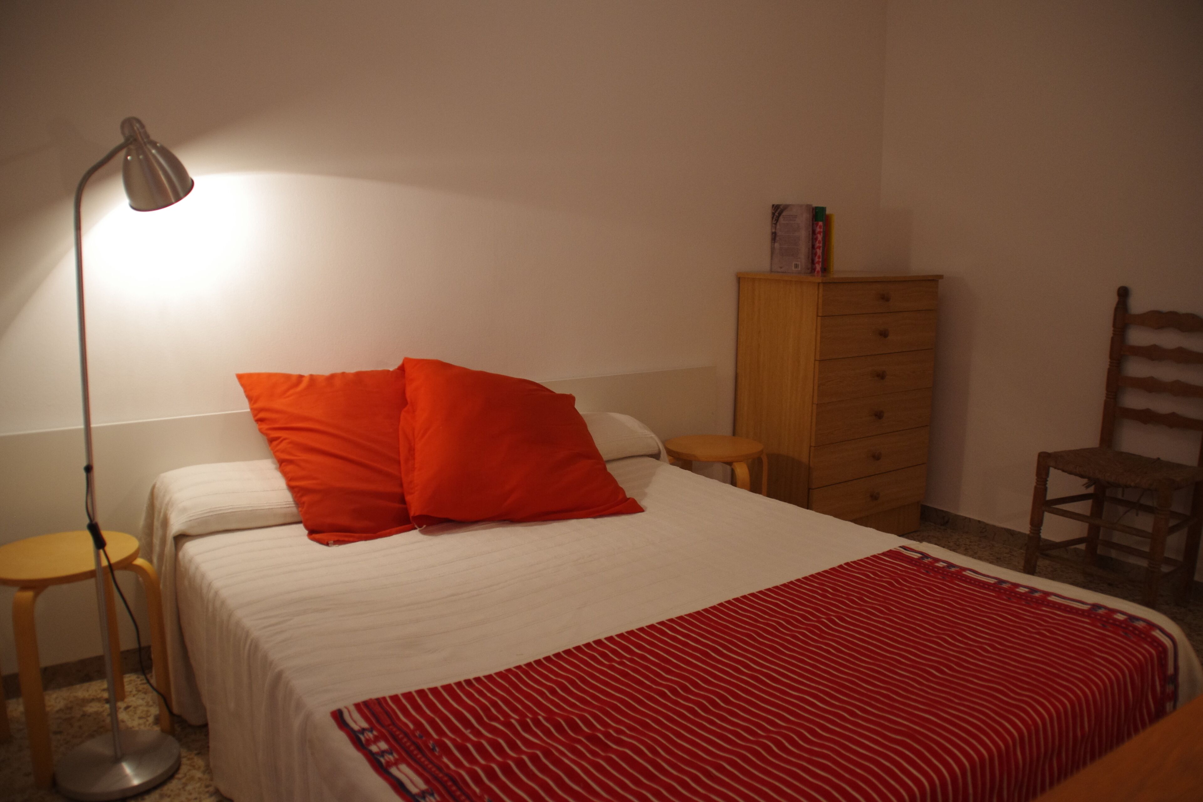 A bedroom with a bed, a red and white striped blanket, a wooden dresser, a chair, and a floor lamp.
