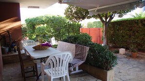 An outdoor patio with a stone floor, a white table, two white chairs, a wicker basket, and a cushioned bench.