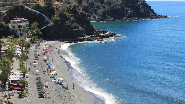 Une plage côtière avec une colline rocheuse, une mer d’un bleu limpide, ainsi que quelques parasols et quelques personnes.