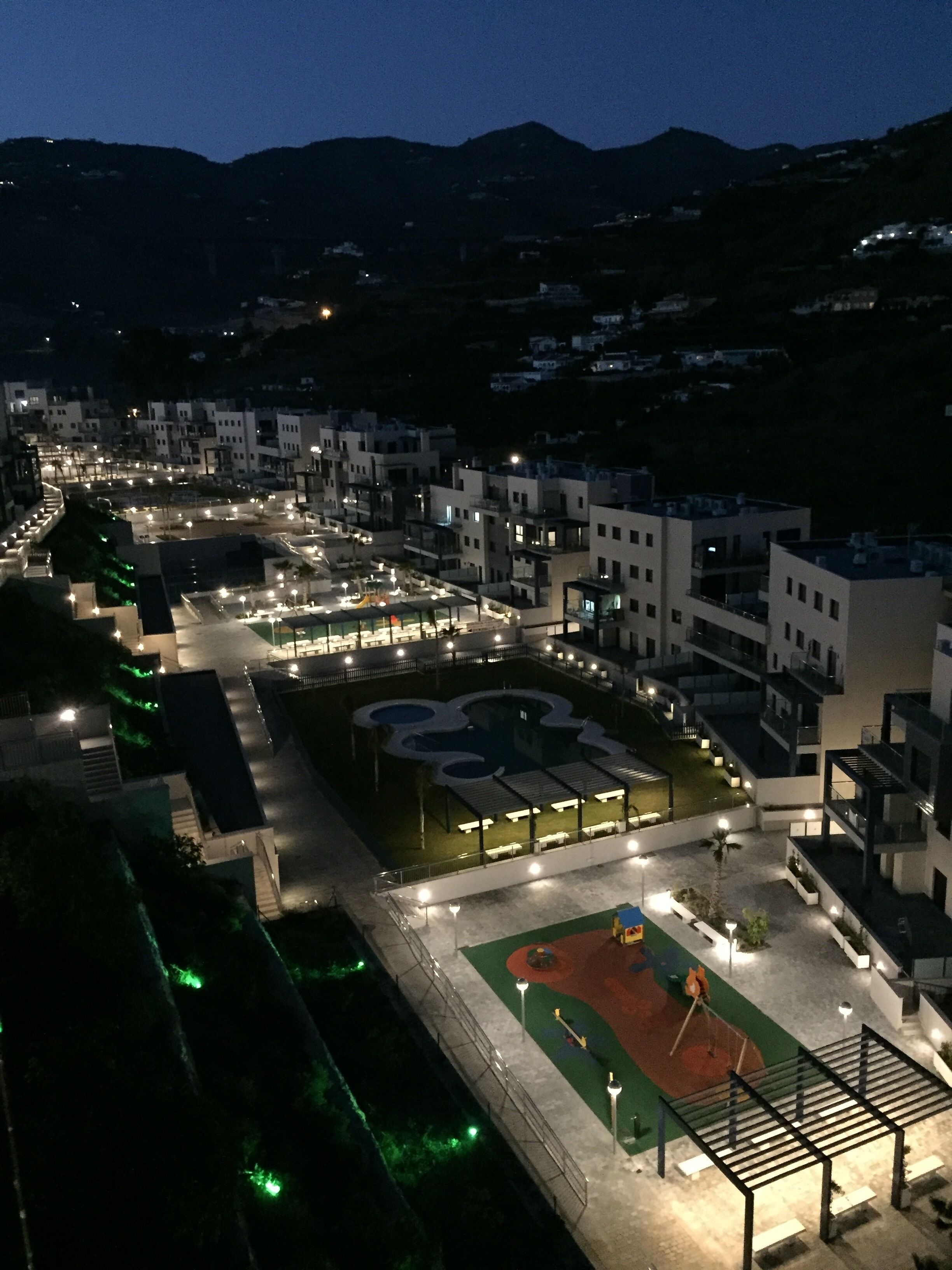 An aerial view of a residential area with modern buildings, a well-lit central square, and a sports field.