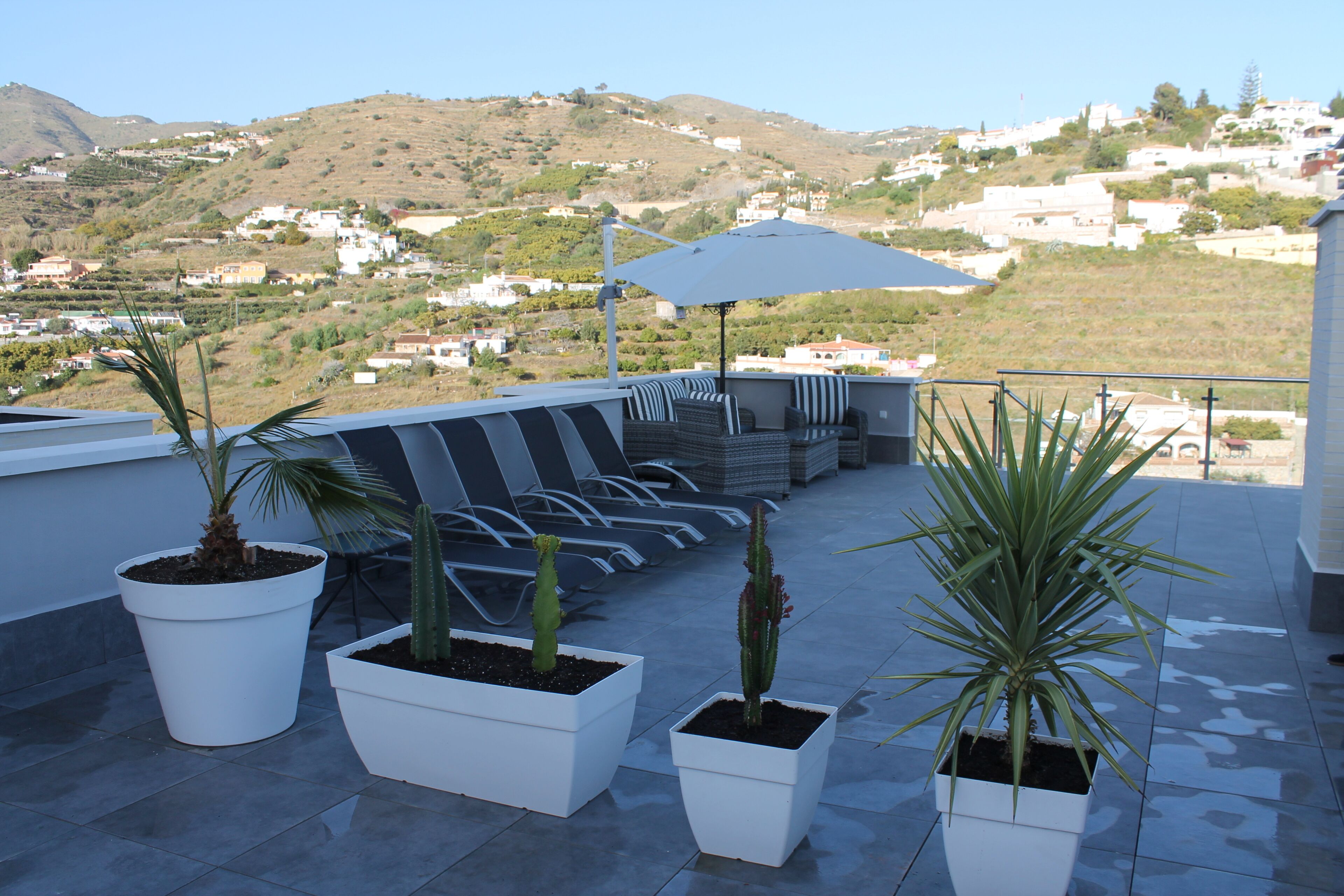 A terrace with potted plants, outdoor seating, and a view of a hillside town.