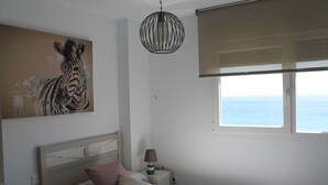 Une chambre à coucher avec un lit, une table de chevet, une lampe, un tableau représentant une zèbre et une fenêtre donnant sur la mer.