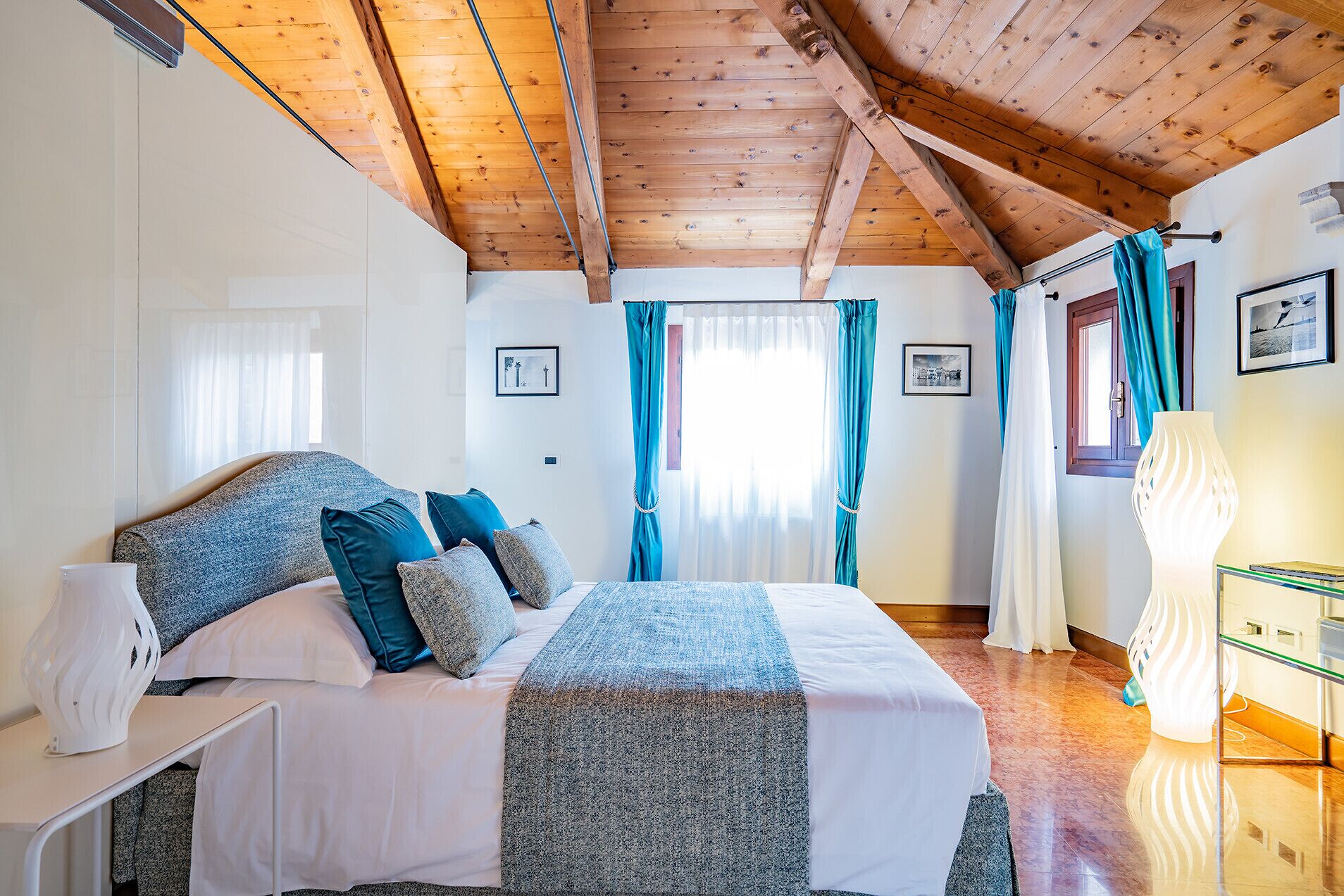 Une chambre avec un plafond en bois, un lit avec une literie blanche et bleue, une table de chevet, une lampe et une fenêtre avec des rideaux.