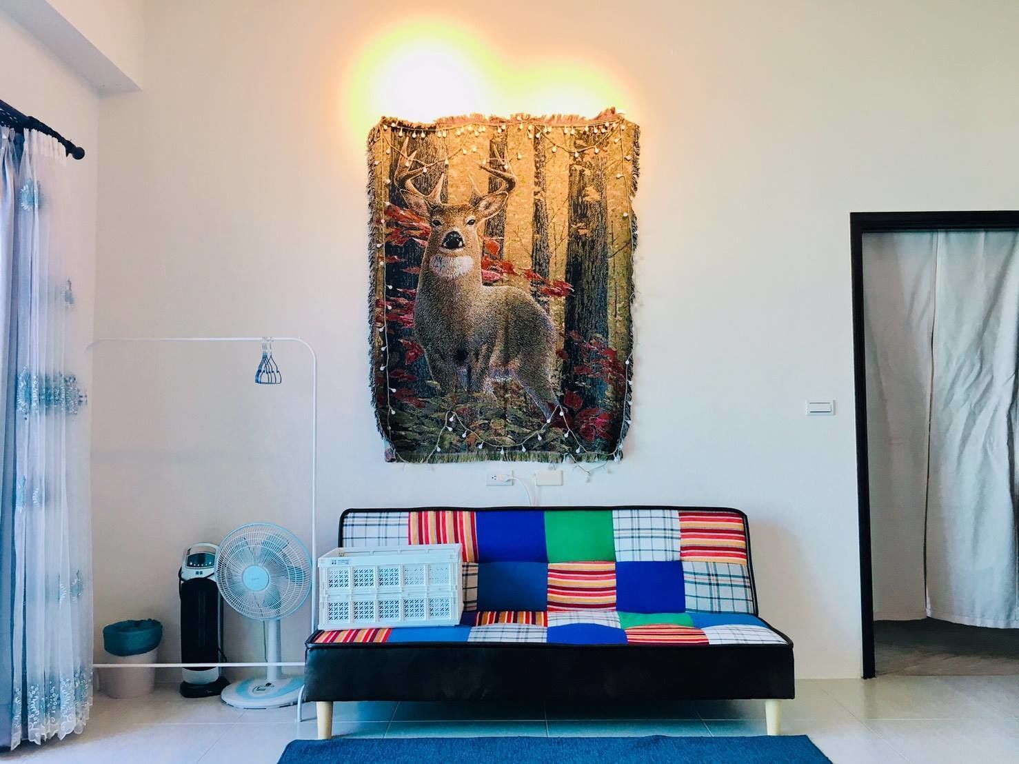 A room with a sofa bed featuring a colorful patchwork design, a wall-mounted tapestry of a deer, a standing fan, and a radiator.