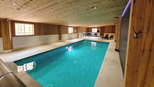 Una piscina interna con pareti e soffitto in legno, un'area salotto con sedie e un tavolo, e una finestra che offre luce naturale.
