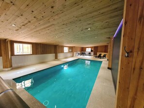 An indoor swimming pool with wooden walls and ceiling, a seating area with chairs and a table, and a window providing natural light. - Country Cottage with Indoor heated swimming pool and panoramic views. (Hope Valley)