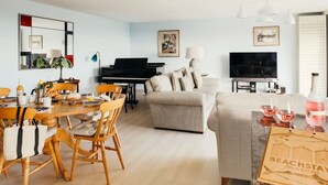 Moderni olohuone, jossa on ruokailutila, piano, sohva, televisio ja juomien kanssa tarjoiluun tarkoitettu pöytä.