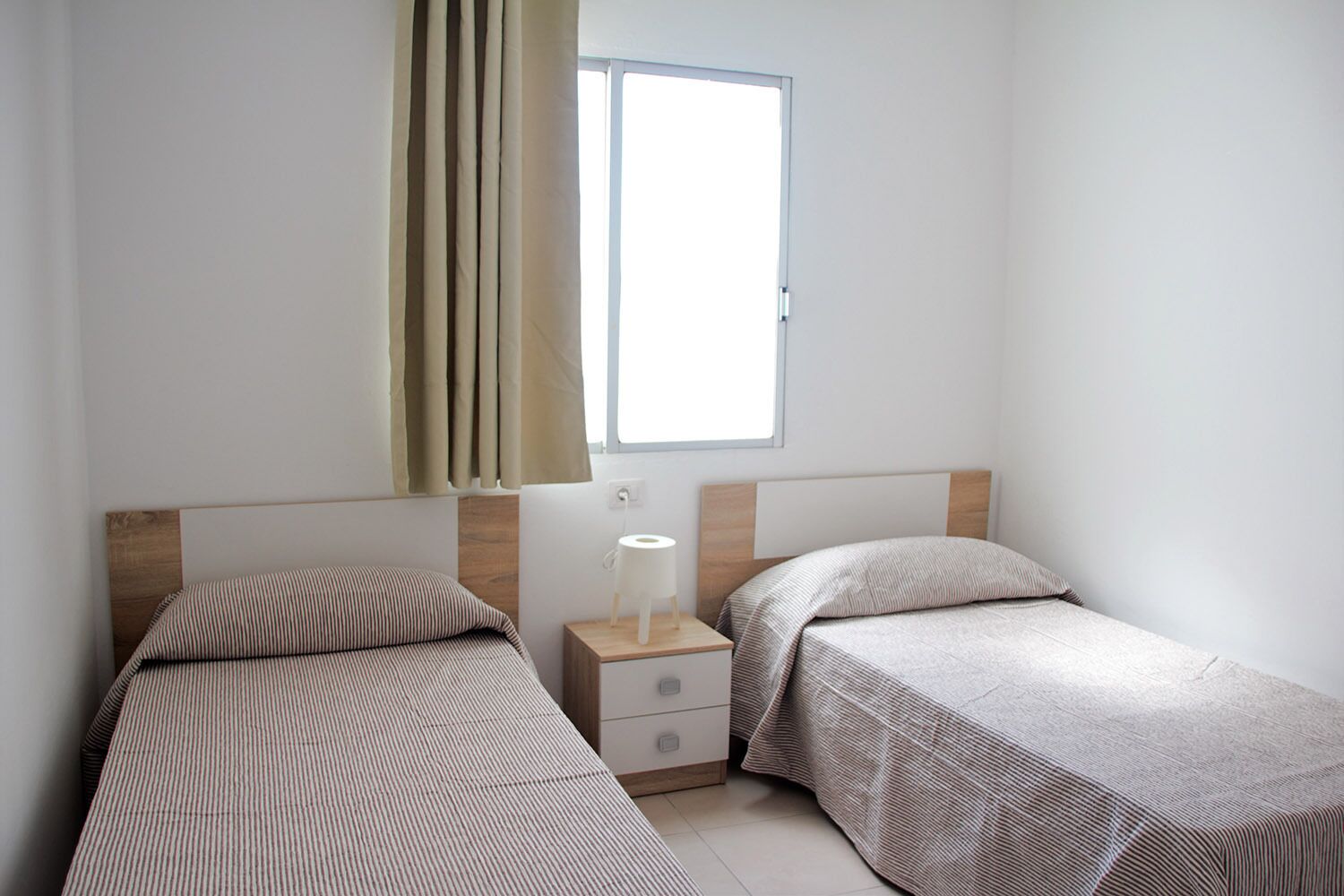 A small, clean room with two single beds, a wooden nightstand with a lamp, and a window with curtains.
