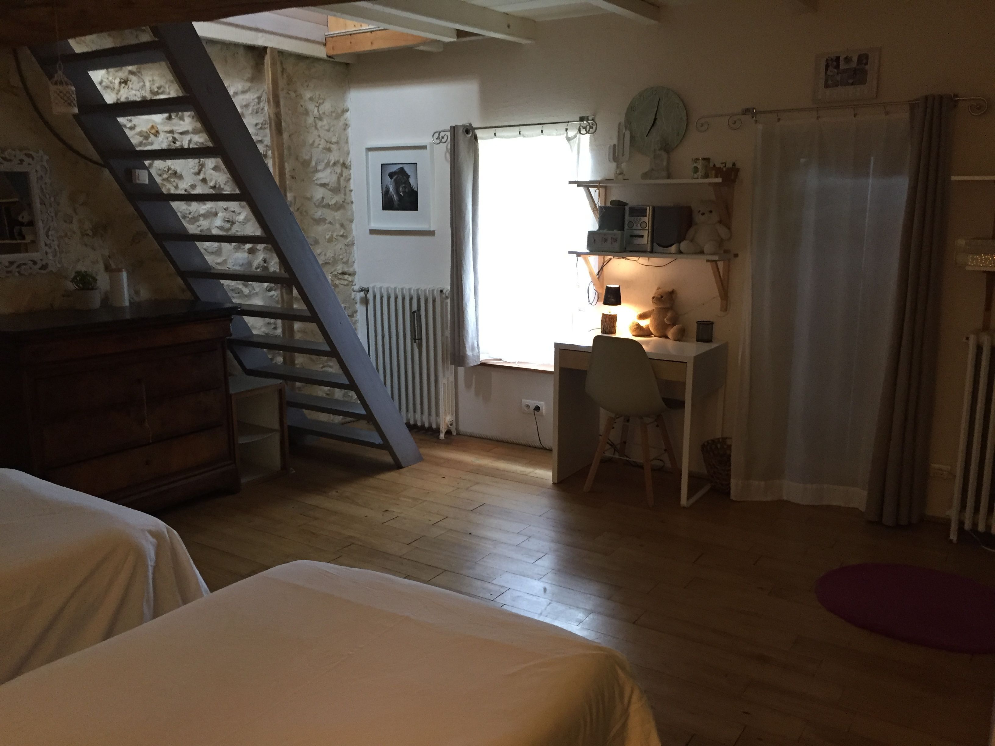 A room with a wooden floor, a desk with a chair, a bed, a staircase, and a radiator.