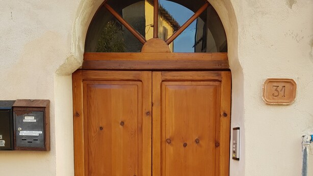 Una puerta doble de madera con un pequeño ventanal arriba, un buzón y una placa con un número en la pared.