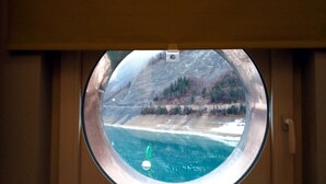 Une vue à travers une fenêtre circulaire, donnant sur une étendue d’eau bordée d’une rive et entourée d’arbres.