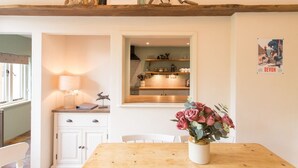 A cozy kitchen with wooden shelves, a dining table with a vase of flowers, and a window with a view of the kitchen. - The Linhay, Kings Nympton - Quiet, very rural thatched barn near great pub. (Kings Nympton)
