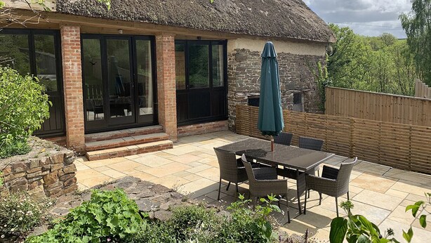 A house with a thatched roof, brick walls, and large windows. There is a patio with a table and chairs, and a wooden fence in the background. - The Linhay, Kings Nympton - Quiet, very rural thatched barn near great pub. (Kings Nympton)
